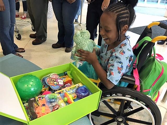 A young child in a wheelchair smiles as she opens a bright green Cheeriodicals box of toys at Ronald McDonald House following a team building event