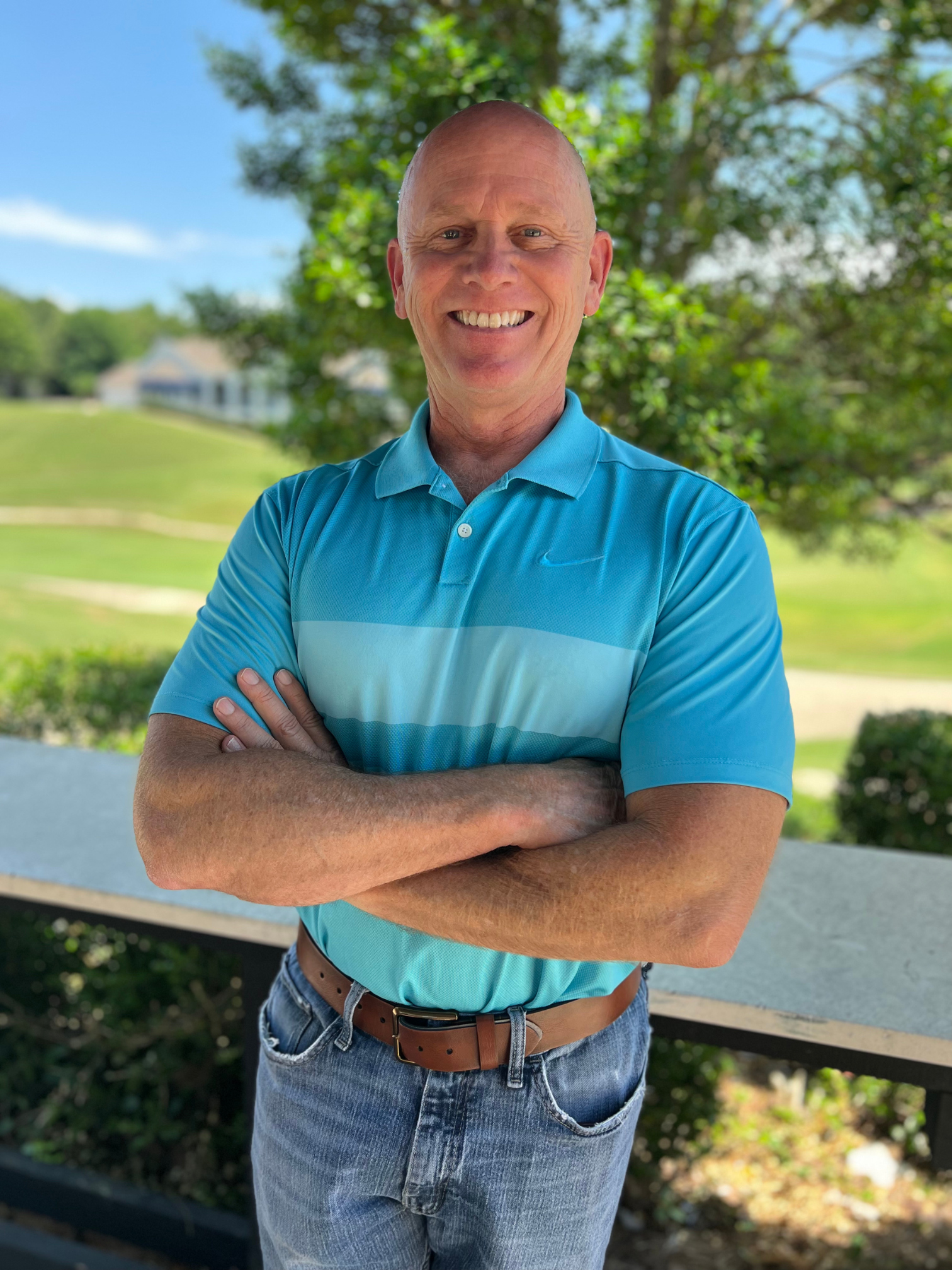 A man smiling at the camera with his arms folded across his chest. He is wearing a bright blue polo with blue jeans and a brown belt.