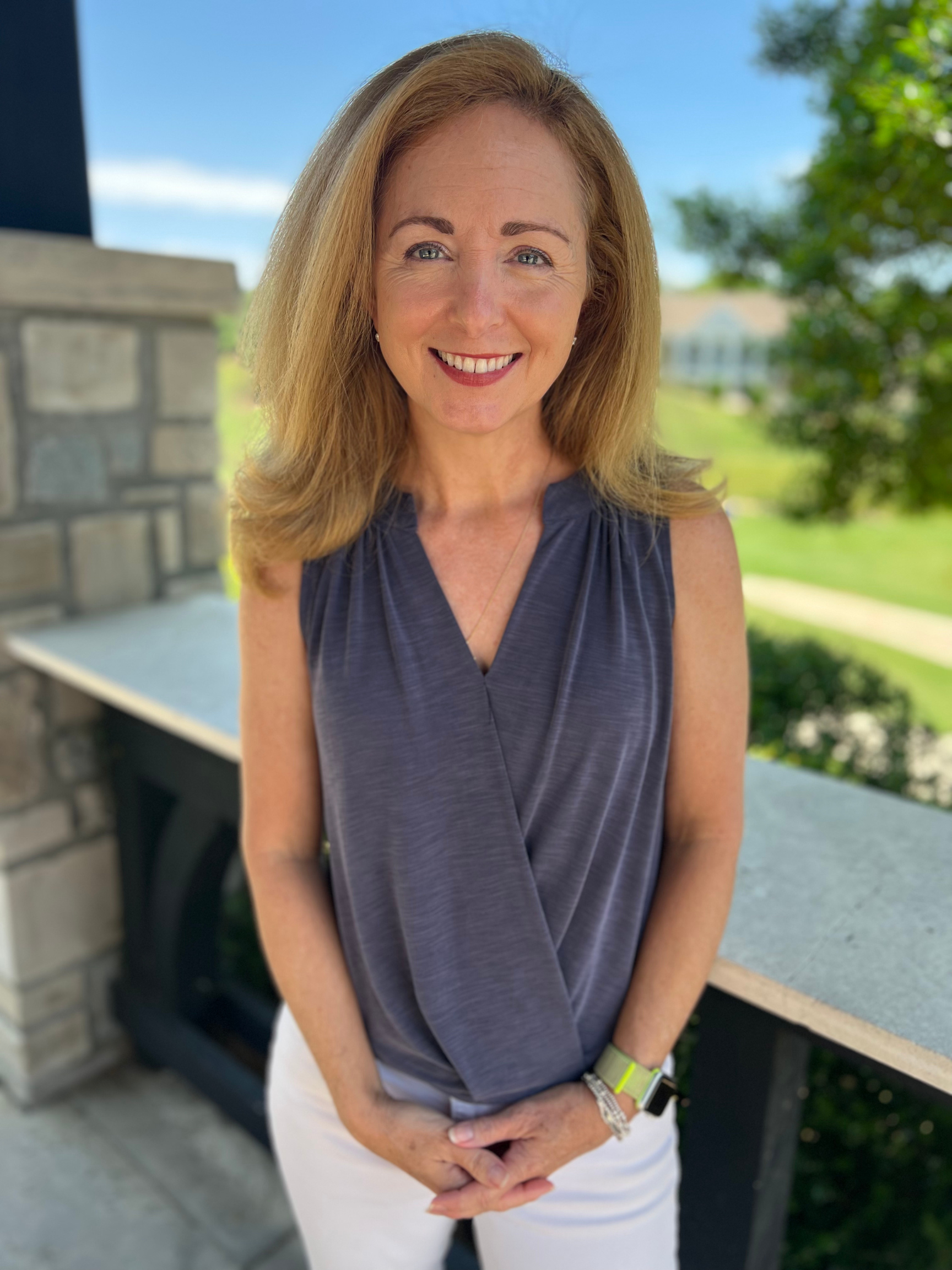 A woman with light blonde hair and blue eyes smiling at the camera. She is wearing a blue tank top and white pants with her hands folded in front of her lower body.