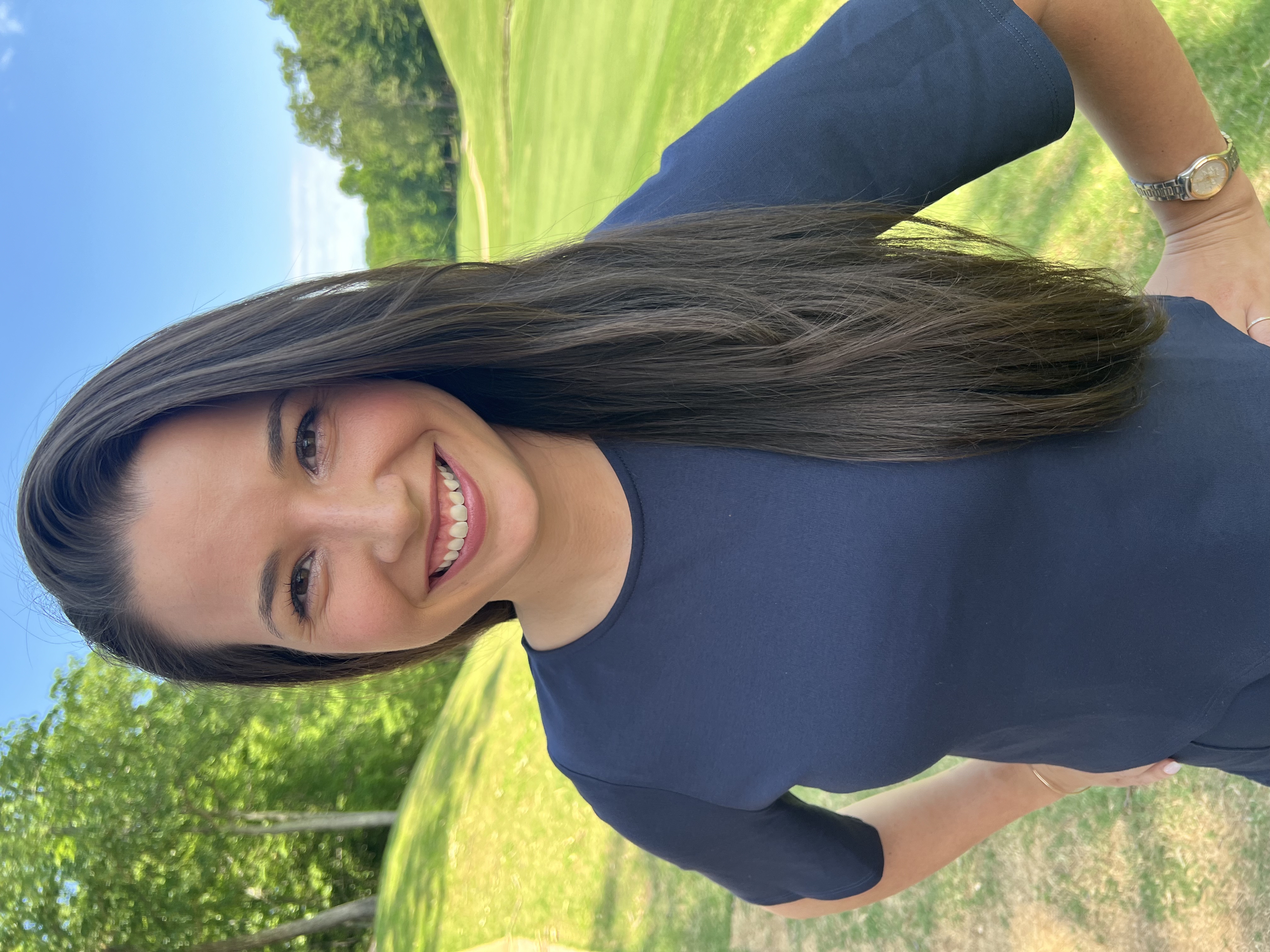 A woman smiling at the camera with long, dark brown hair and brown eyes.  She is wearing a navy blue dress and has her hands on her hips.  There is bright green grass behind her. 