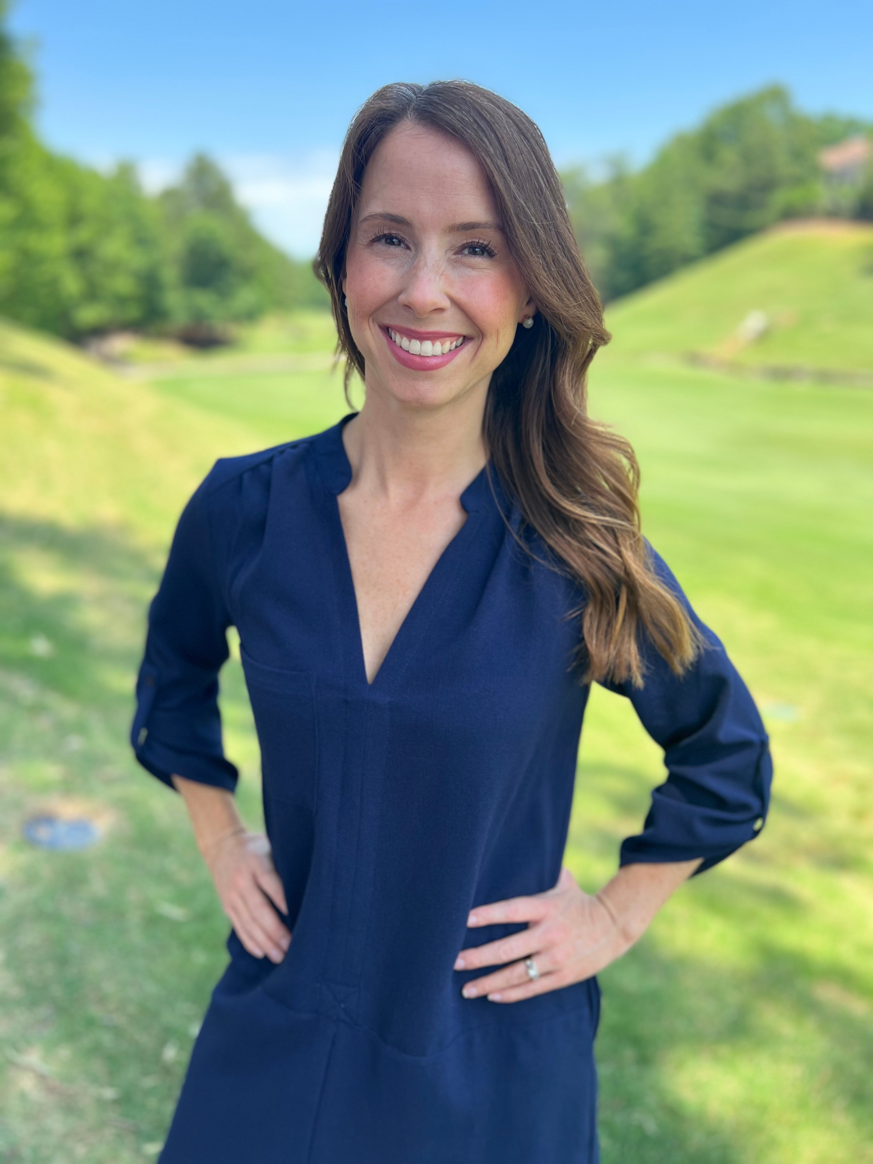 A woman smiling with long brown hair and a blue dress with her hands on her hips. There is bright green grass behind her.