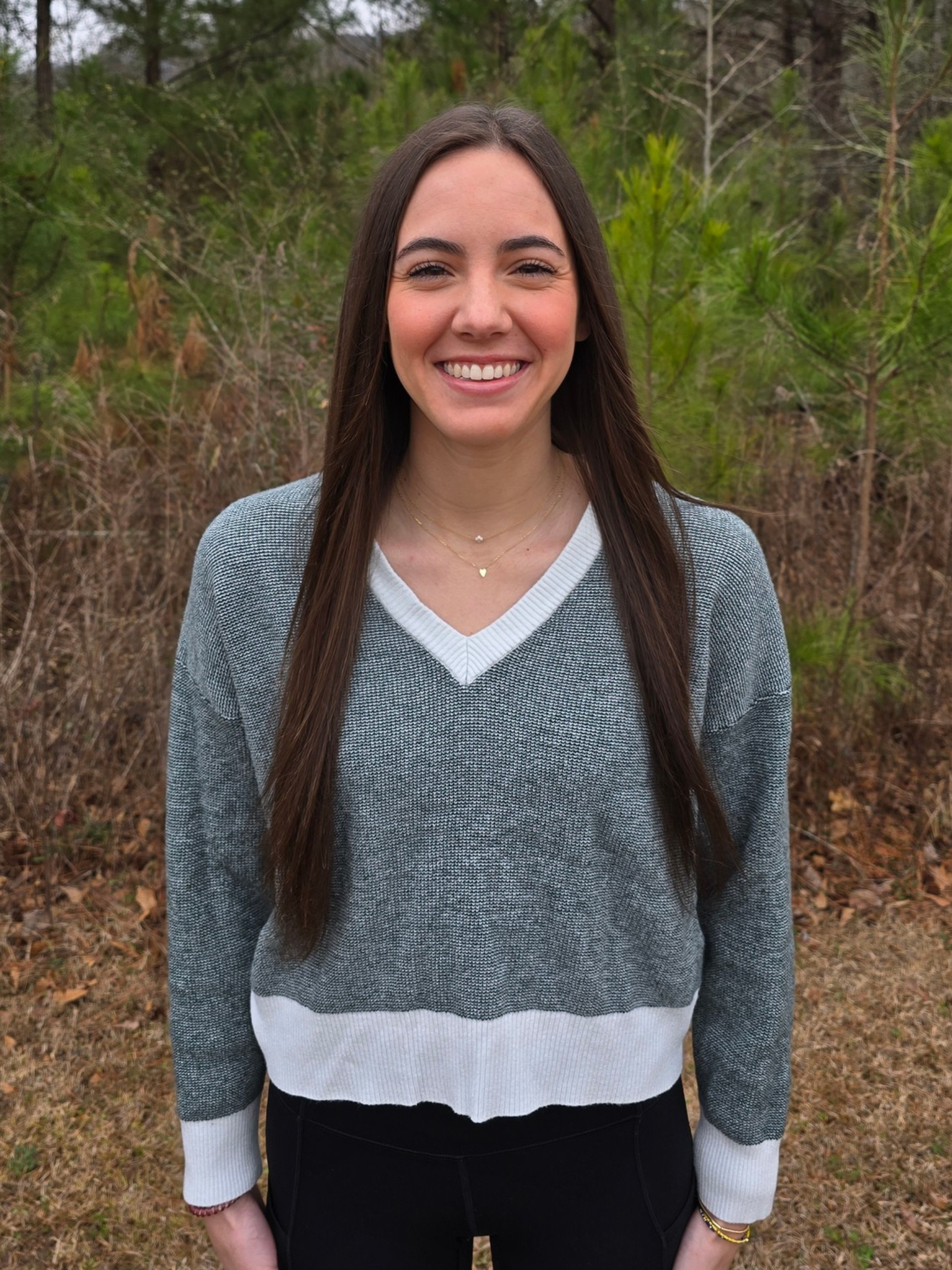 A woman smiling with long light brown hair in a green dress wearing pearl earrings. There is bright green grass behind her and a stone pillar.
