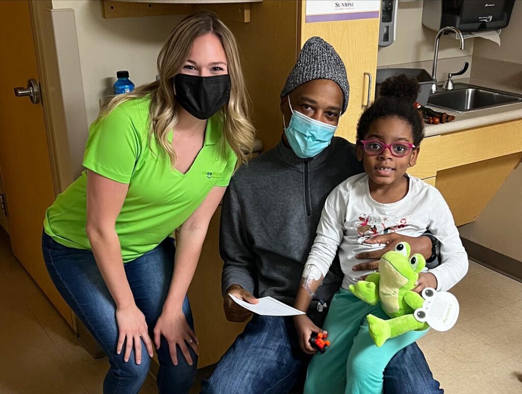 A Cheeriodicals staff member smiles and poses with a little girl and her mom in the hospital