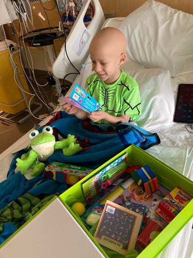 A little boy sitting in a hospital bed looks through his new bright green Cheeriodicals box of toys and smiles