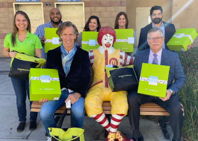 7 San Diego Ronald McDonald House staff members smiles and pose with Ronald McDonald on a bench and are holding bright green Cheeriodicals care boxes and bags for families and kids