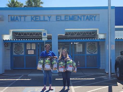A guy and girl stand outside of Matt Kelly Elementary School holding Cheeriodicals backpacks of supplies to donate