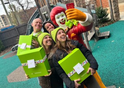 Four people smile and pose with a statue of Ronald McDonald. They are holding bright green Cheeriodicals gift boxes for families at the Ronald McDonald House after a team building event.