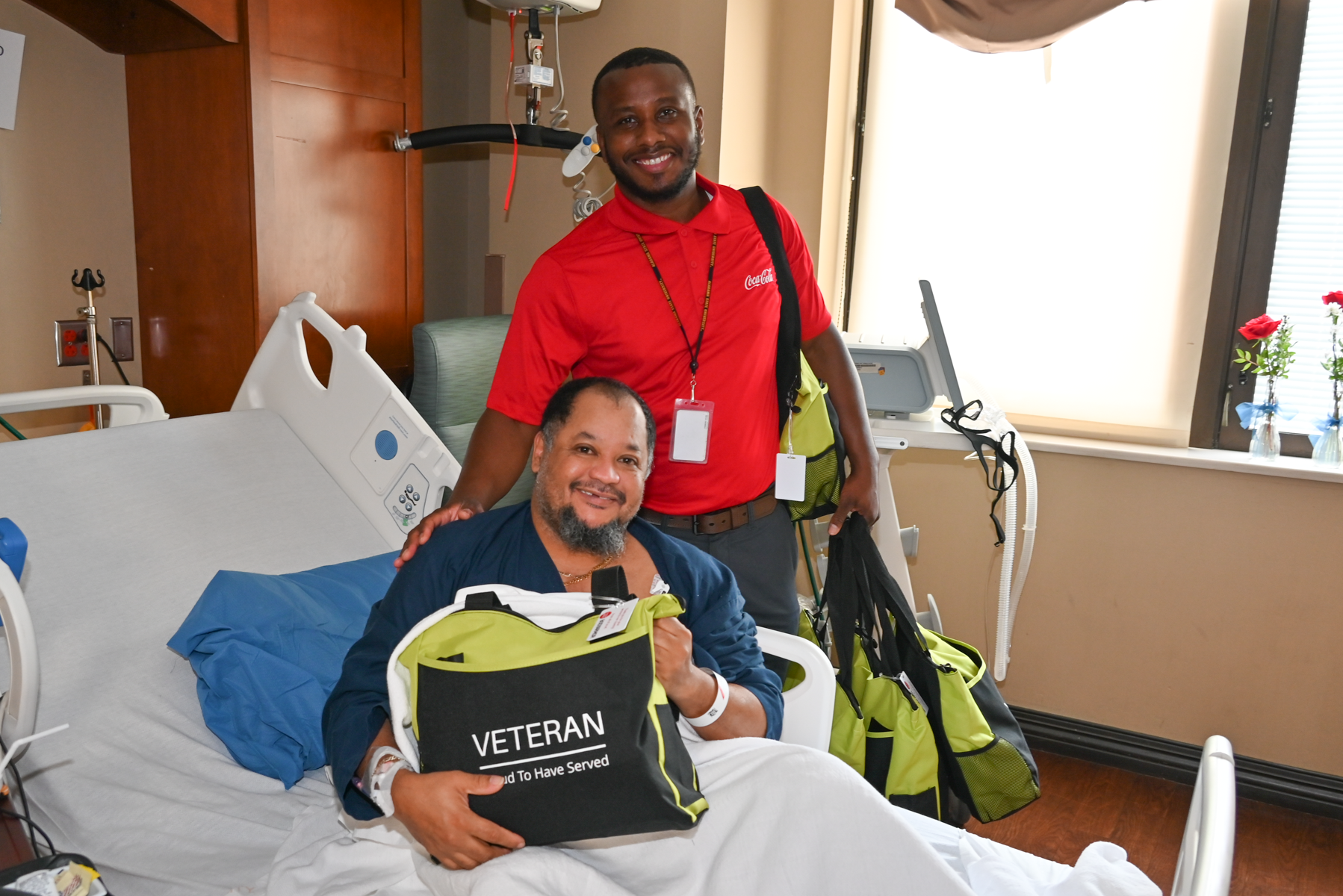 A young boy smiles as he lays in a hospital bed. He is holding a notecard and a green Cheeriodicals box of toys that was packed by a corporate team during a team building activity